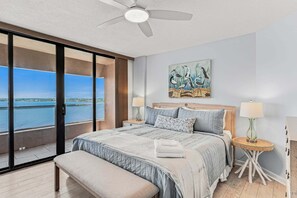 Un dormitorio con una cama grande, una banca, un ventilador de techo, un cuadro y vista al agua.