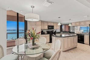 A modern kitchen with a large island, a dining area with glass-top table, wicker chairs, and a view of the sea. - 🌞CBC 1-15A – Water views from every window! 5 balconies, heated pool, pickleball, gym. (Clearwater Beach)