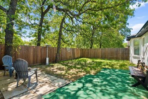 Ein Garten mit Holzlattenzaun, einer Terrasse mit Stühlen, einem grünen Rasen und einem Haus im Hintergrund.