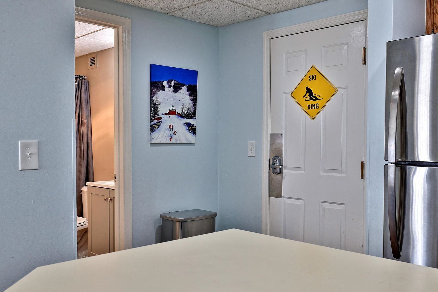 A hotel room with a bed, a door with a ski crossing sign, a painting of a snowy landscape, and a trash bin.