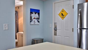 A hotel room with a bed, a door with a ski crossing sign, a painting of a snowy landscape, and a trash bin.
