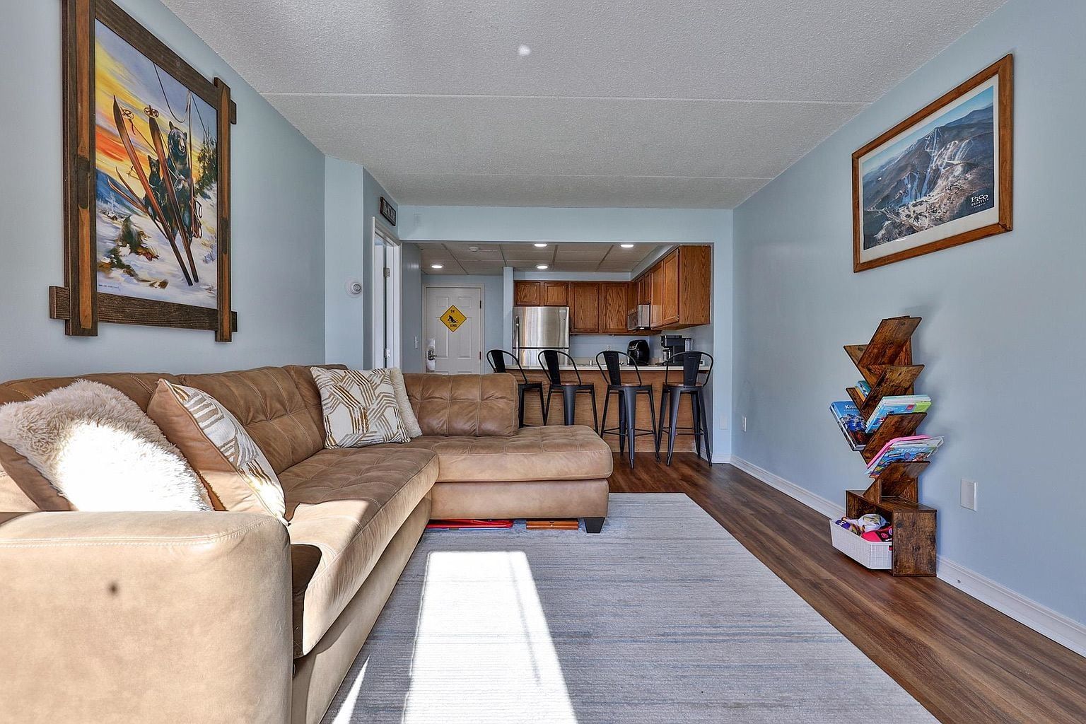 A living room with a sectional sofa, a kitchen in the background, and a bookshelf.