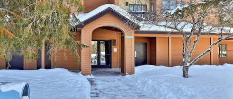 A building with a covered entrance, a snow-covered ground, and a tree in the foreground.