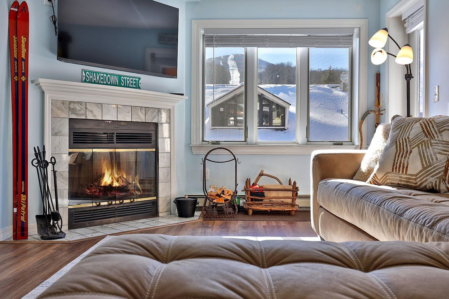 A cozy living room with a fireplace, a television mounted above it, and a large window offering a view of a snowy landscape.
