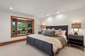 Ein Schlafzimmer mit einem großen Bett, zwei Nachttischen, einem Fenster mit Blick auf Bäume und Einbaustrahler.