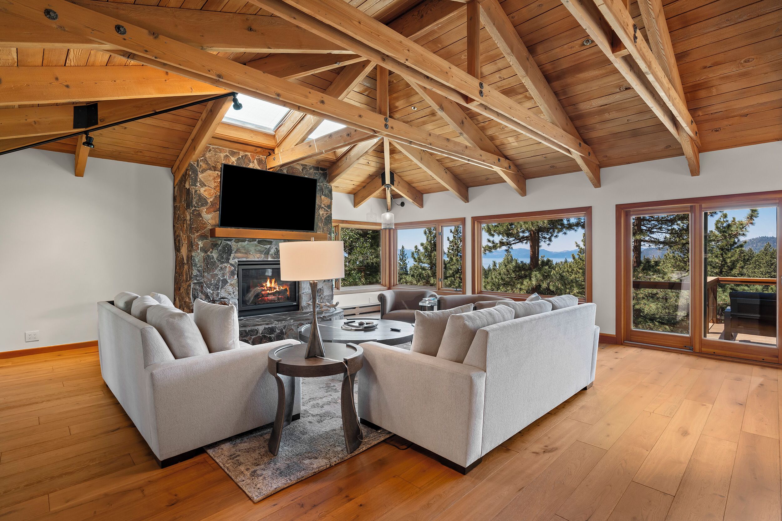 A spacious living room with a stone fireplace, a flat-screen TV, and large windows offering a view of trees and mountains.