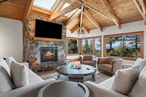 A cozy living room with a stone fireplace, comfortable seating, and large windows offering a view of trees and mountains. - Lake View home 4BR 5BA gym, game room, Large kitchen (Incline Village)