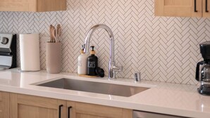 A kitchen with wooden cabinets, a herringbone-patterned backsplash, a white countertop, a stainless steel sink, and a coffee maker.