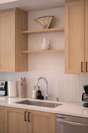 A kitchen with wooden cabinets, a herringbone-patterned backsplash, a white countertop, a stainless steel sink, and a coffee maker. - Refined 1B1B with Pool, Spa and 4K Smart TVs (Montclair)