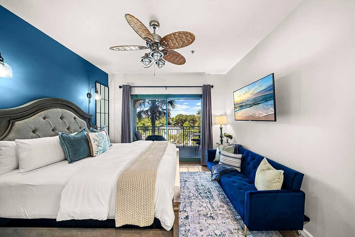 A bedroom with a bed, a blue sofa, a ceiling fan, and a balcony with a view.