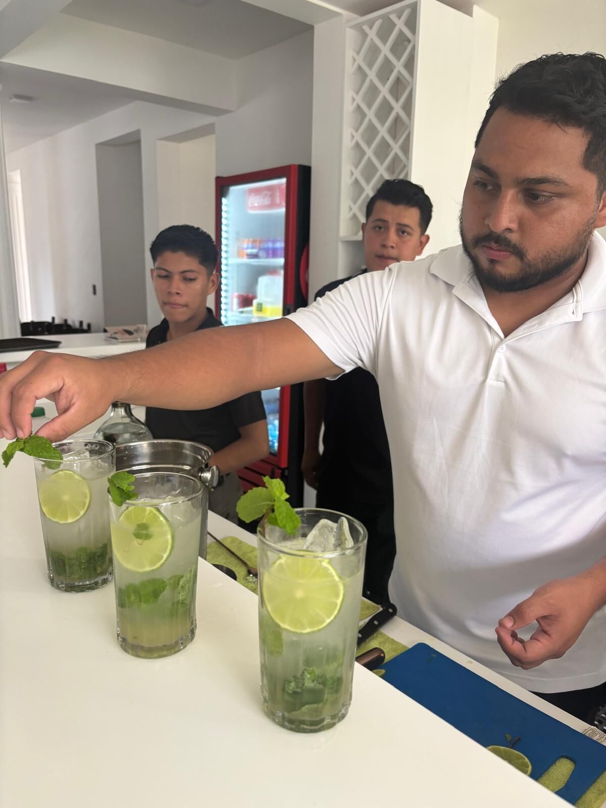 A bar area with a bartender preparing drinks, a refrigerator in the background, and a person standing behind the bar.