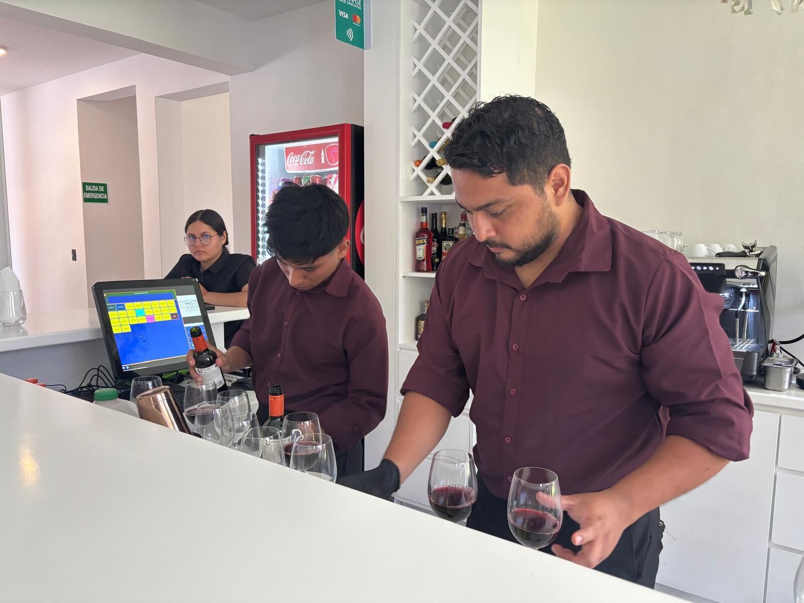 A bar area with two servers attending to customers, a Coca-Cola refrigerator, and a coffee machine.