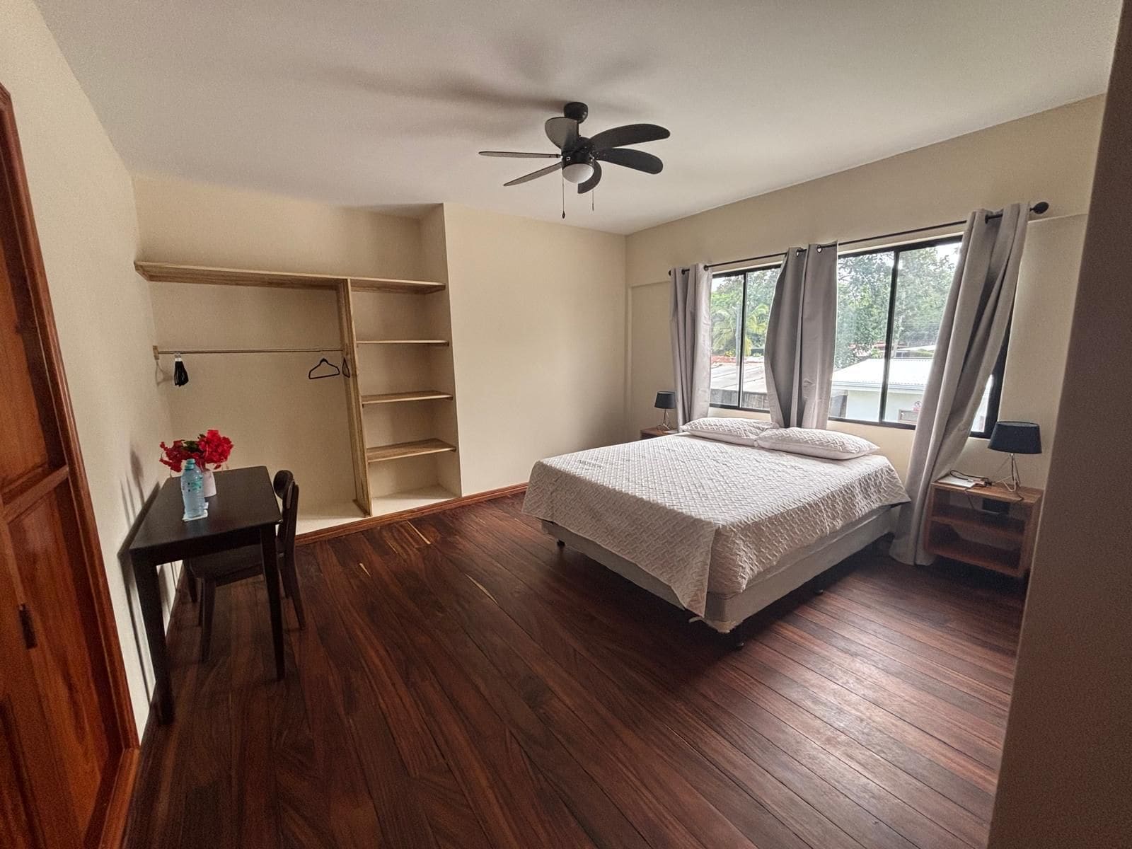 Un dormitorio con cama, ventilador de techo, ventana con cortinas, mesita de noche y suelo de madera.