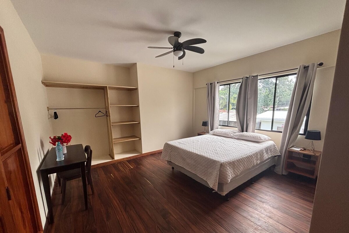 A bedroom with a bed, a ceiling fan, a window with curtains, a bedside table, and a wooden floor.