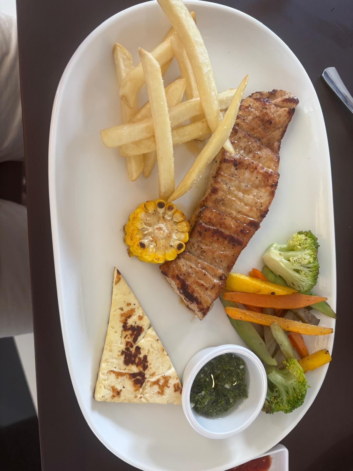 A plate with grilled meat, French fries, grilled corn, grilled vegetables, a piece of flatbread, and a small bowl of sauce.