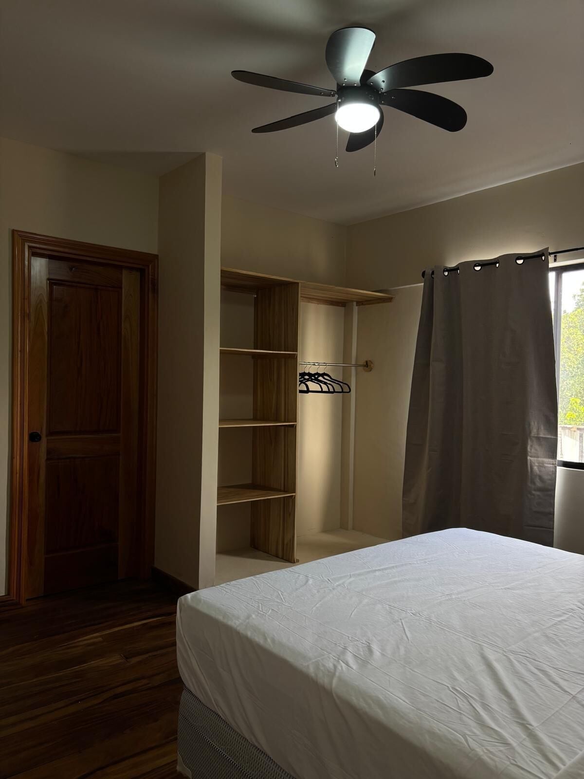 Quarto com cama, ventoinha no teto, guarda-roupa de madeira e janela com cortinas.