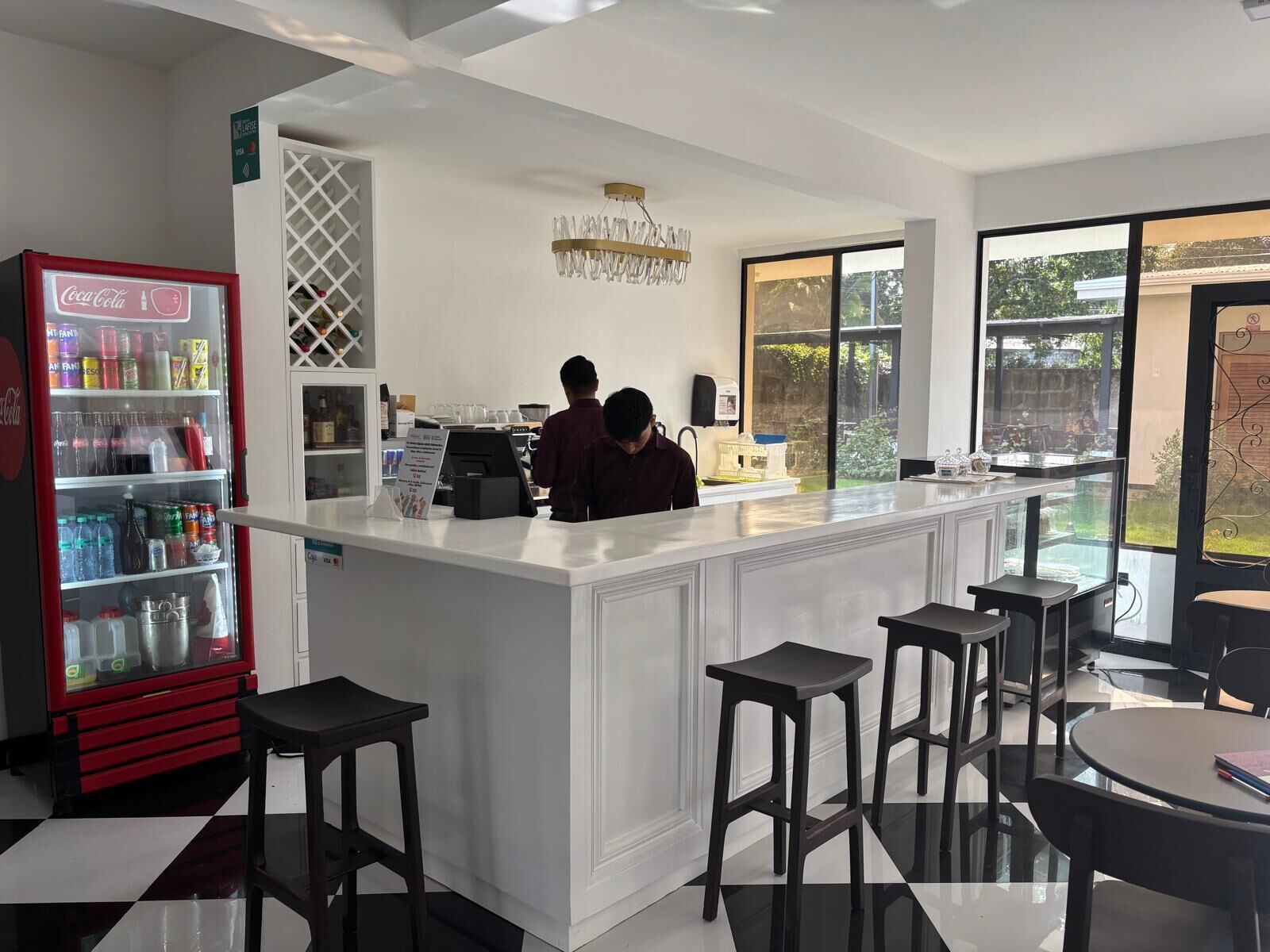Zona de bar con mostrador blanco, taburetes, refrigerador de Coca-Cola y un ventanal con vistas al exterior.