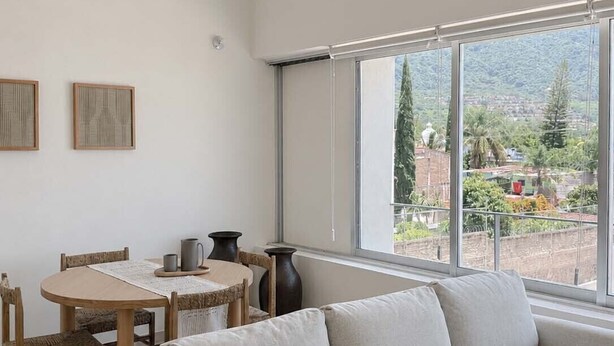 A modern living room with a sofa, a dining table with chairs, and a large window offering a view of greenery and buildings.