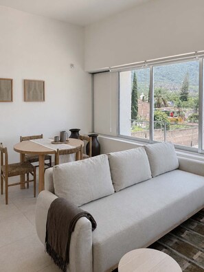 A modern living room with a sofa, a dining table with chairs, and a large window offering a view of greenery and buildings.