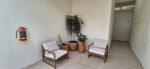A small outdoor seating area with two wooden chairs, a fire extinguisher mounted on the wall, and two potted plants.