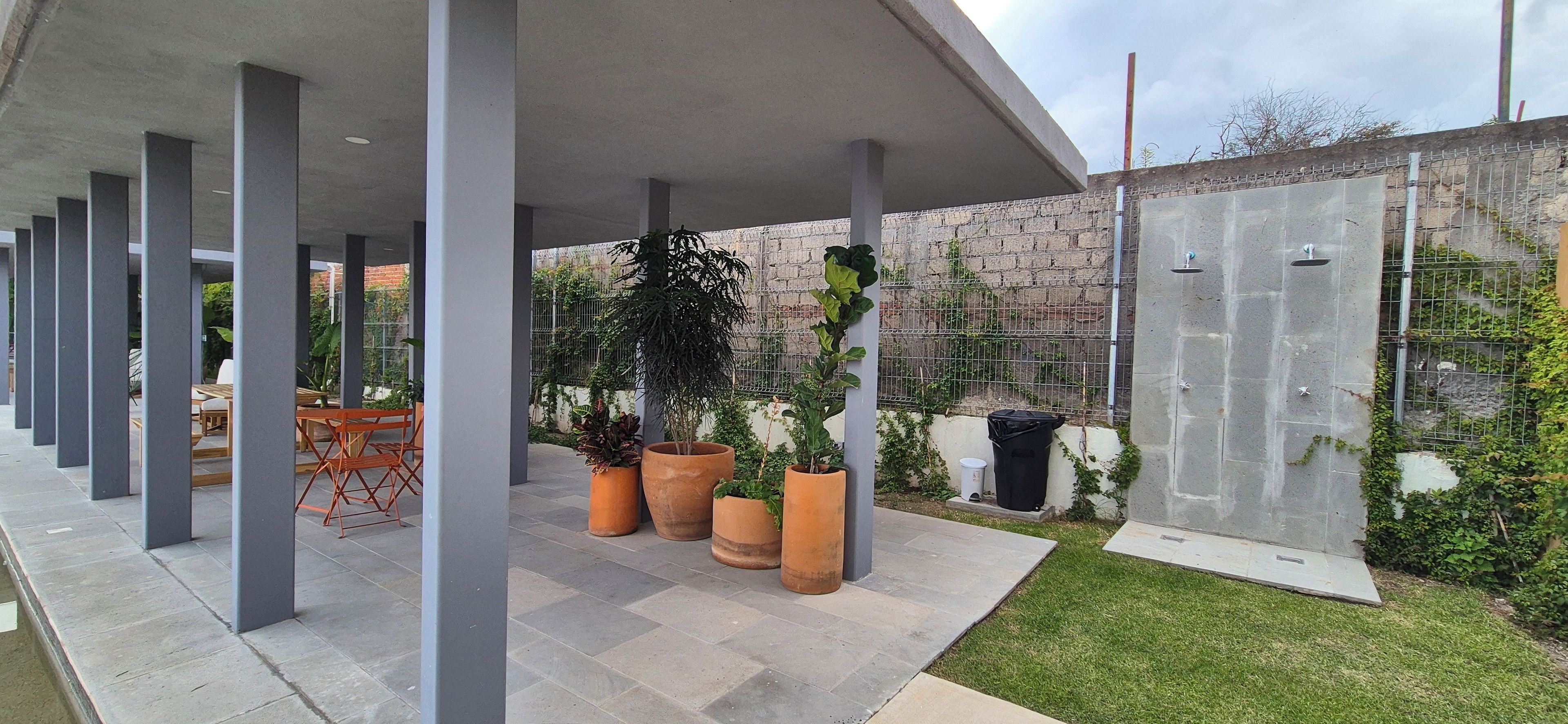 A covered outdoor area with a row of chairs and tables, potted plants, and a stone wall with a metal fence.