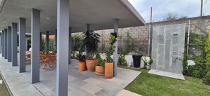 A covered outdoor area with a row of chairs and tables, potted plants, and a stone wall with a metal fence.