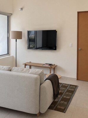 A modern living room with a sofa, a flat-screen TV mounted on the wall, a lamp, a small wooden table, and a door.