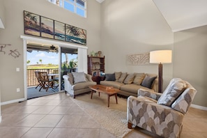 A spacious living room with a high ceiling, a sliding glass door leading to an outdoor area, and a large window providing natural light.