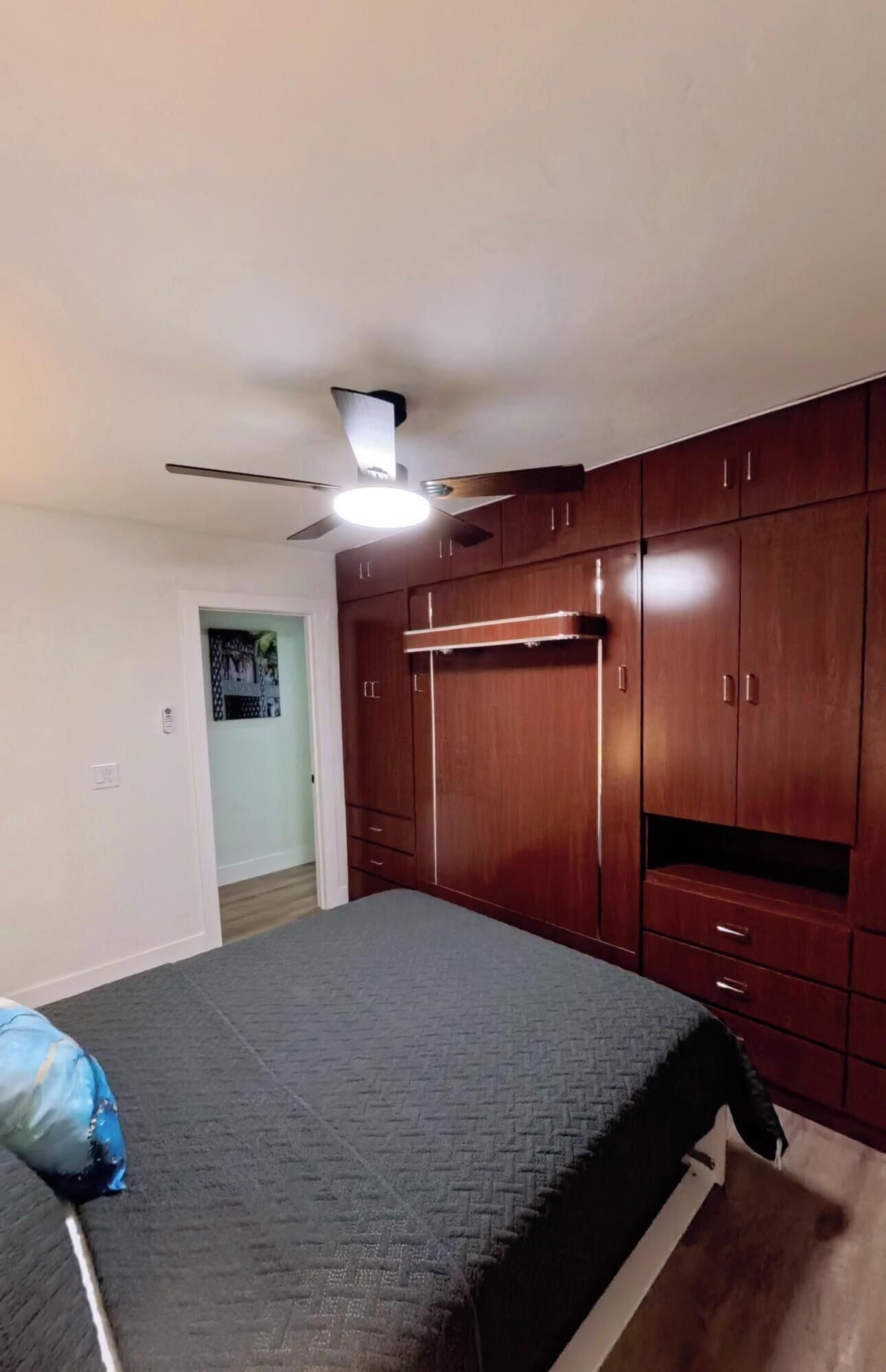 A bedroom with a bed, wardrobe, ceiling fan, and a door leading to another room.