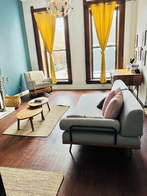 Ein Wohnzimmer mit einer Couch, einem Couchtisch, einem Sessel und einem Schreibtisch mit Lampe und Büchern. Zwei Fenster mit Vorhängen, eine Chandelier und eine Wand mit gerahmten Bildern.