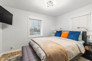 Ein Schlafzimmer mit Bett, Nachttisch, an der Wand montiertem Fernseher und einem Fenster mit Jalousien.
