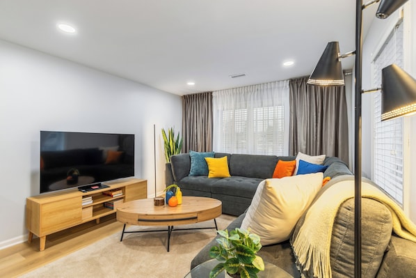 Ein modernes Wohnzimmer mit einem Flachbildfernseher, einem Holztisch, einem grauen Sofa mit bunten Kissen und einem großen Fenster mit Vorhängen.
