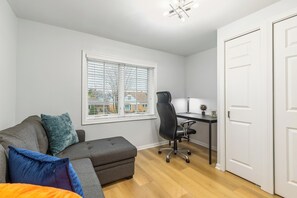 Une petite pièce bien éclairée, avec un canapé gris, un fauteuil de bureau noir, une petite table avec une lampe et une fenêtre avec des stores.