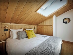 A bedroom with a sloped ceiling, wooden paneling, a skylight, a bed with white bedding, a grey knitted throw, and a round mirror on the wall. - Penthouse Mont Grange - Enjoy the mountain views at any time - OVO Network (La Chapelle-d'Abondance)