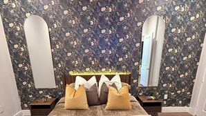 A bedroom with a floral wallpaper, a large bed with a central headboard, two nightstands, and a mirror on one wall.
