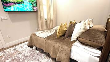 A bedroom with a bed, a television mounted on the wall, a book on a side table, and a window with curtains.