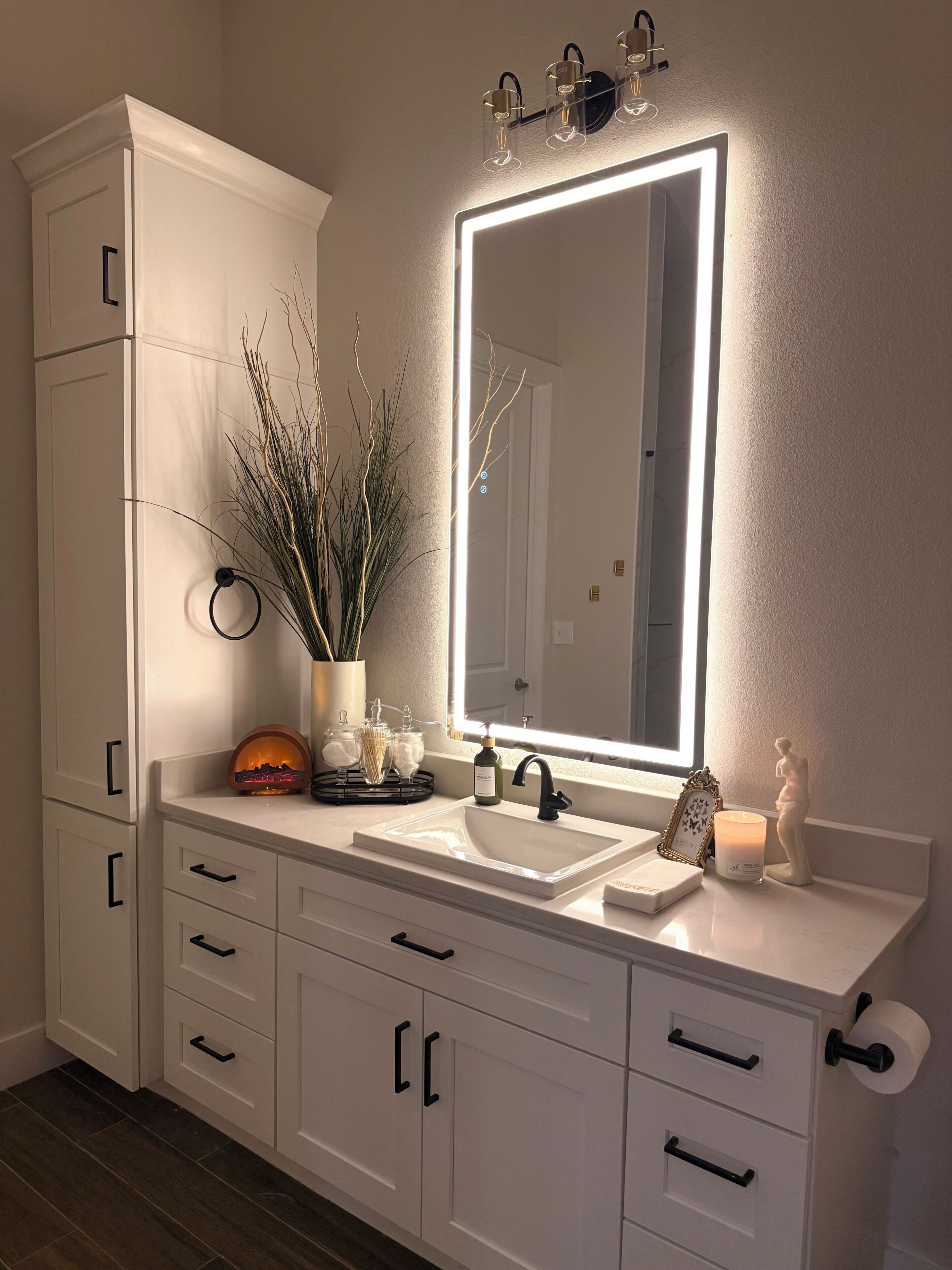 Une salle de bain avec un meuble-lavabo blanc, un miroir avec éclairage intégré, un lavabo et une plante décorative.
