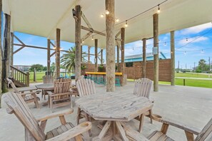 An outdoor patio with wooden furniture, a covered area with a white roof, and a view of a building in the background. - Bolivar 5BR Beach House - Fenced - Steps to Beach (Bolivar Peninsula)