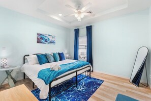 Ein Schlafzimmer mit einem Bett, einem Deckenventilator, einem Fenster mit Vorhängen, einem Gemälde an der Wand und einem kleinen Tisch mit einer Lampe.