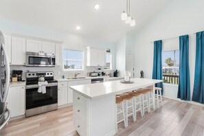 A modern kitchen with white cabinetry, stainless steel appliances, a central island with stools, and a view through a window with blue curtains. - Bolivar 5BR Beach House - Fenced - Steps to Beach (Bolivar Peninsula)