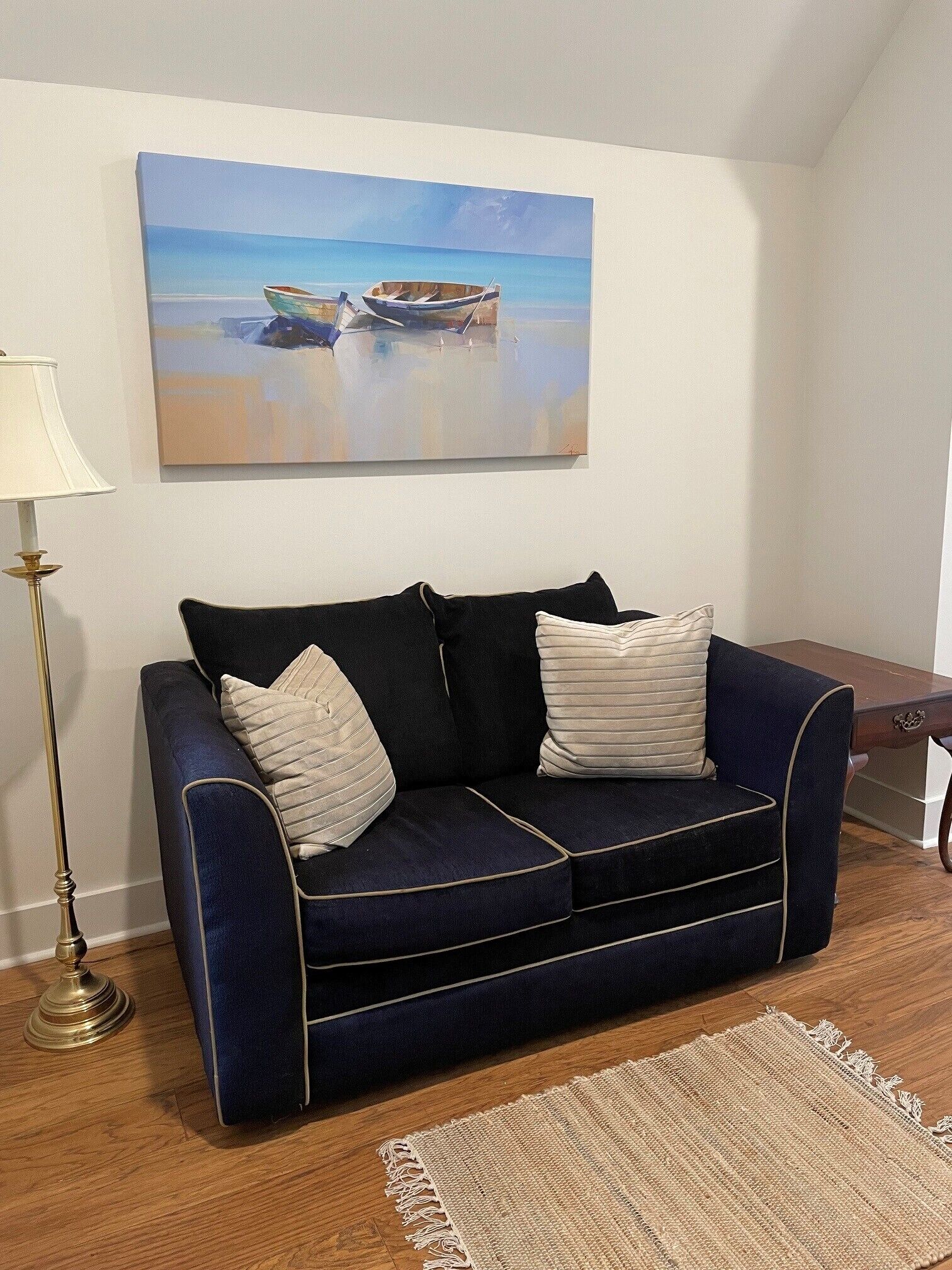 Ein dunkelblaues Sofa mit zwei Kissen, einer Lampe und einem Gemälde von Booten an einem Strand.
