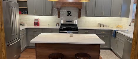 A kitchen with grey cabinets, a central island with stools, a stove, and pendant lighting.