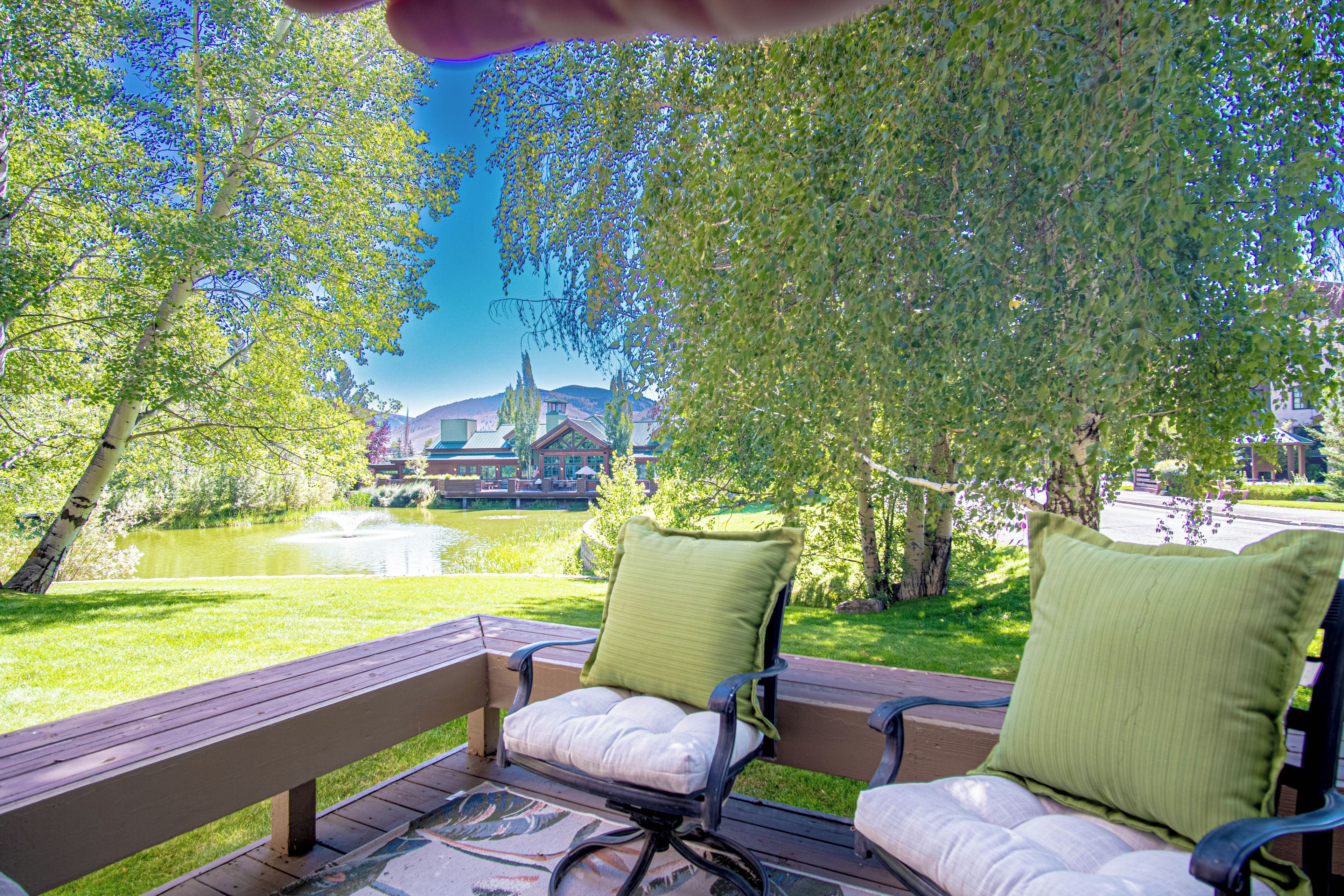 An outdoor seating area with two chairs and a table, overlooking a pond and a building with a tower.