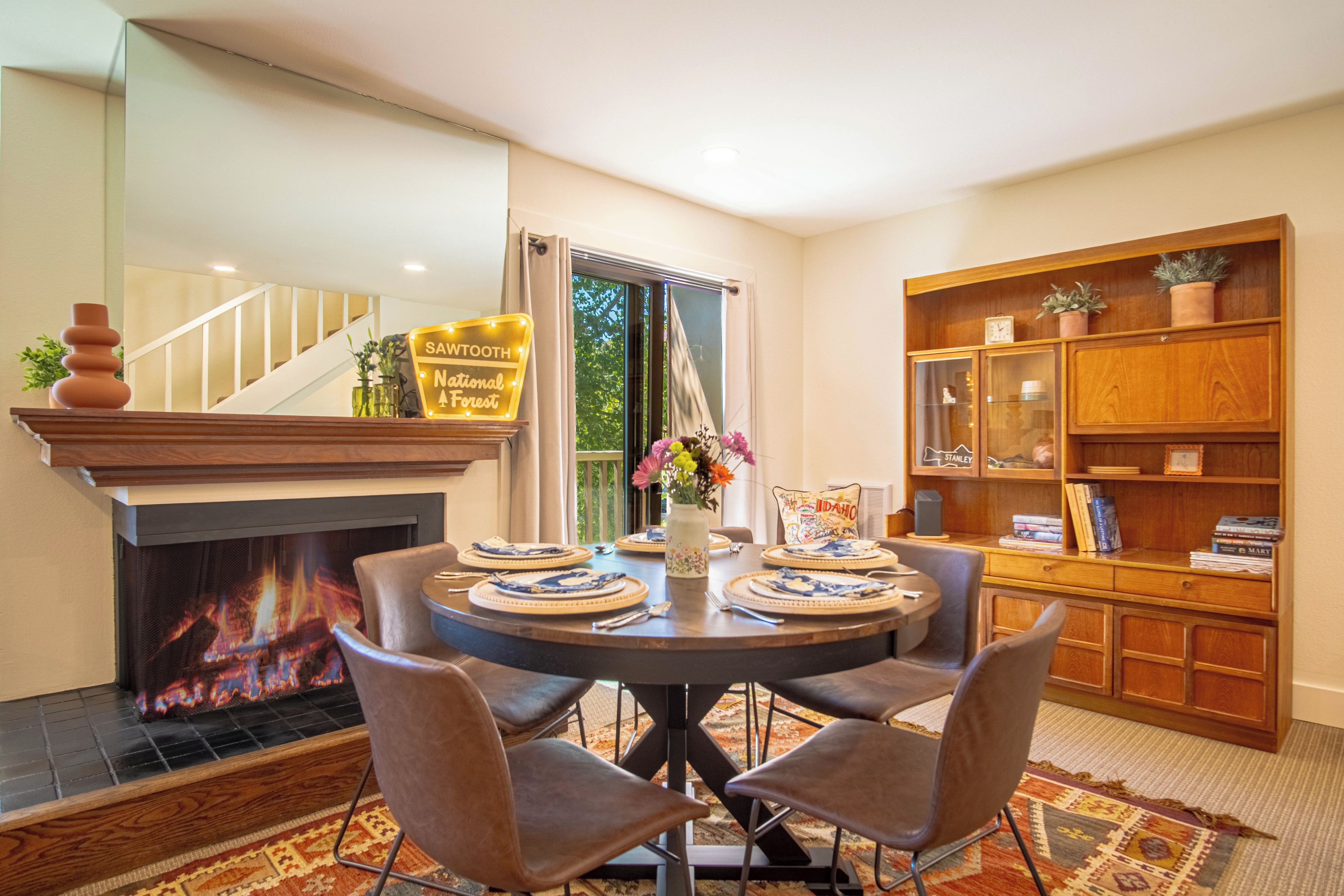 Uma sala de jantar com lareira, uma mesa redonda posta para quatro pessoas e uma estante com livros e objetos decorativos.