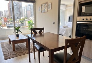 A hotel room with a dining table, chairs, a potted plant, a bed with white linens, and a view of the cityscape through large windows. - Edificio Eleuterio by Domus Travels (Santiago)