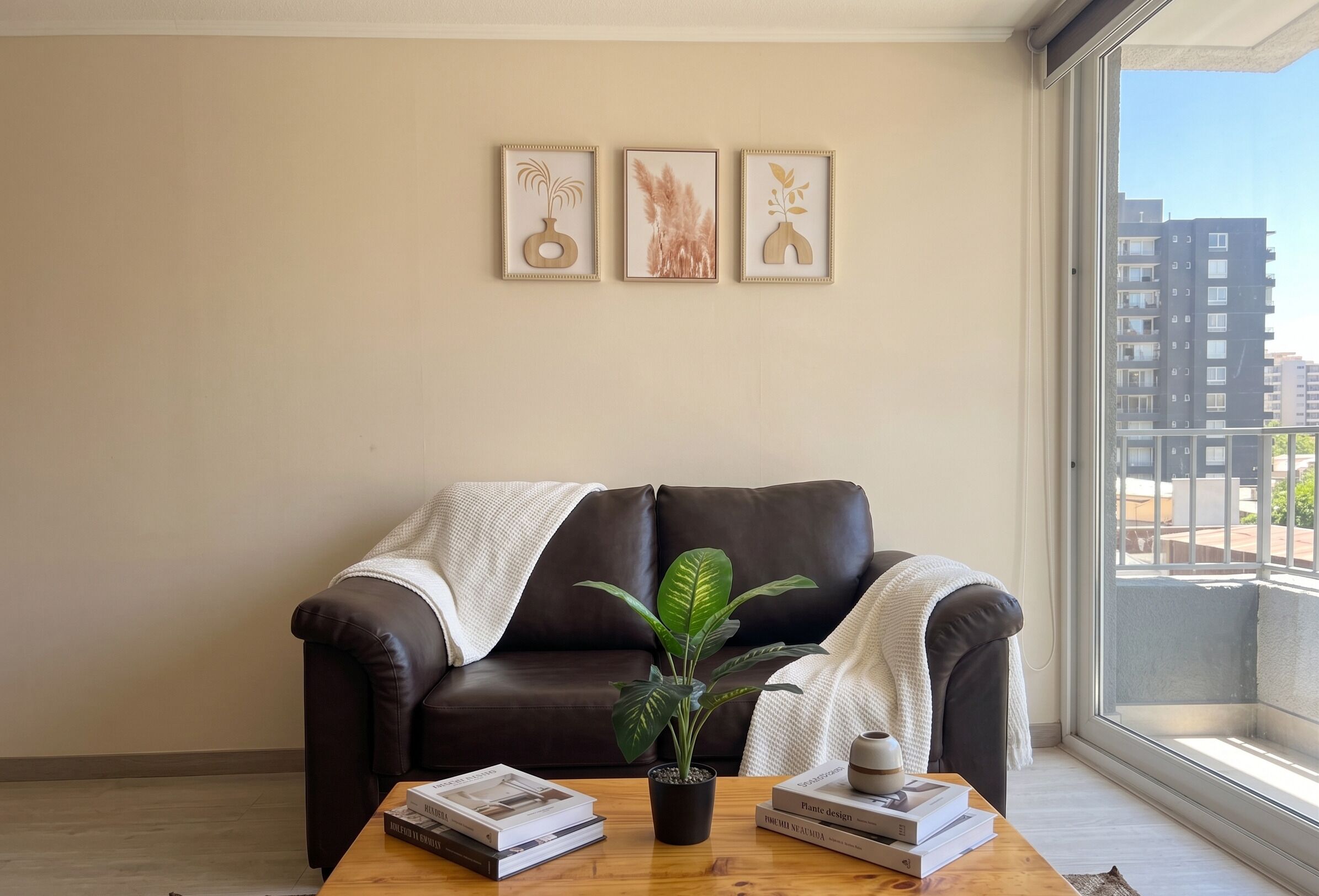Un salón con un sofá de cuero marrón, una mesa de centro con libros y una planta, y un ventanal con vista a la ciudad.