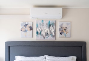 Ein Schlafzimmer mit grauem Kopfteil, einem weißen Bett und drei abstrakten Bildern an der Wand.