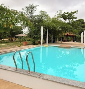A swimming pool with a clear blue water surface, surrounded by greenery and a gazebo structure in the background.
