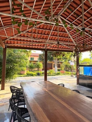 An outdoor covered seating area with a long wooden table and chairs, surrounded by greenery and a building in the background.