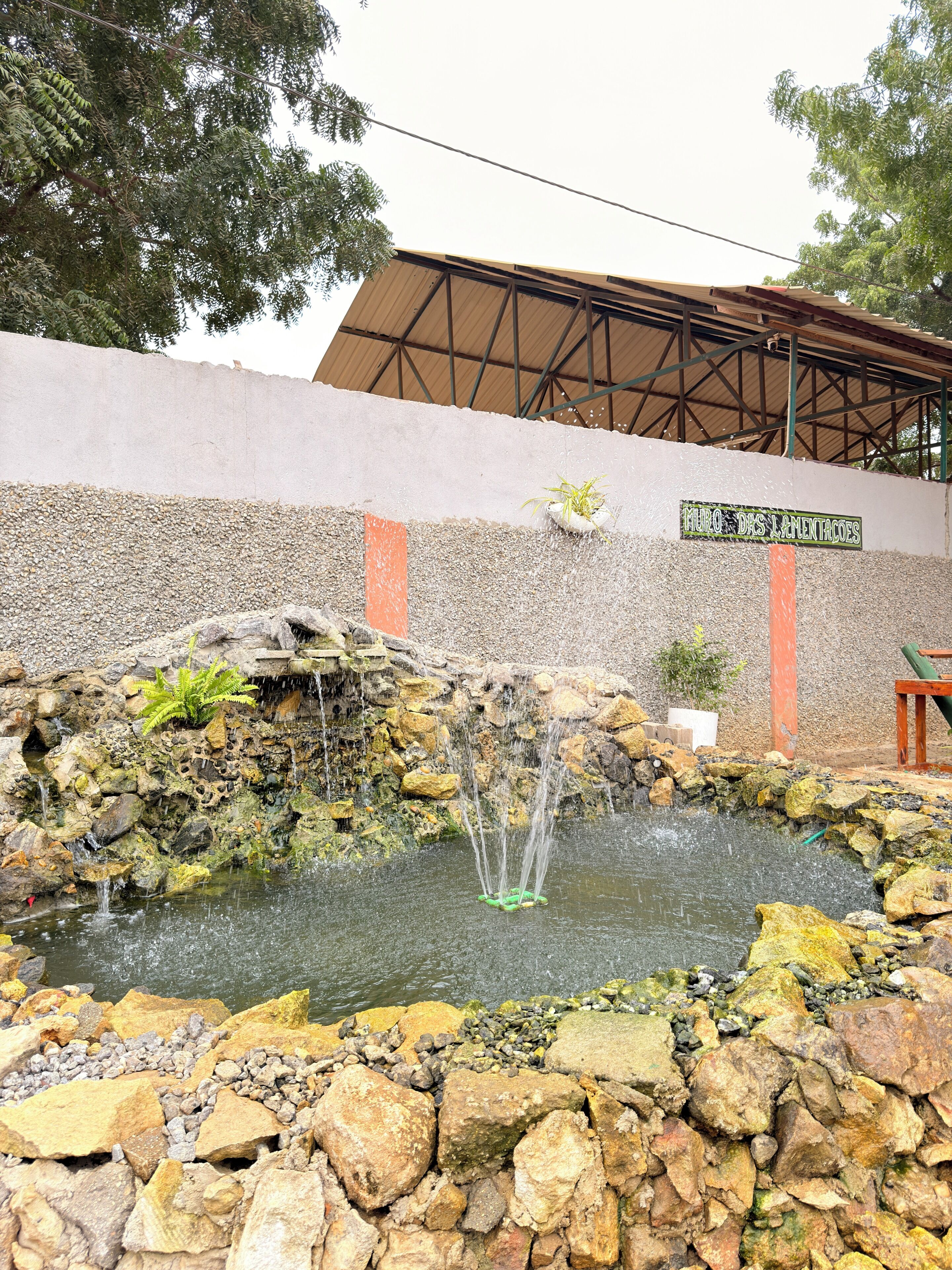 Um elemento aquático com uma fonte, rodeado por pedras e plantas, com um edifício e um letreiro ao fundo.